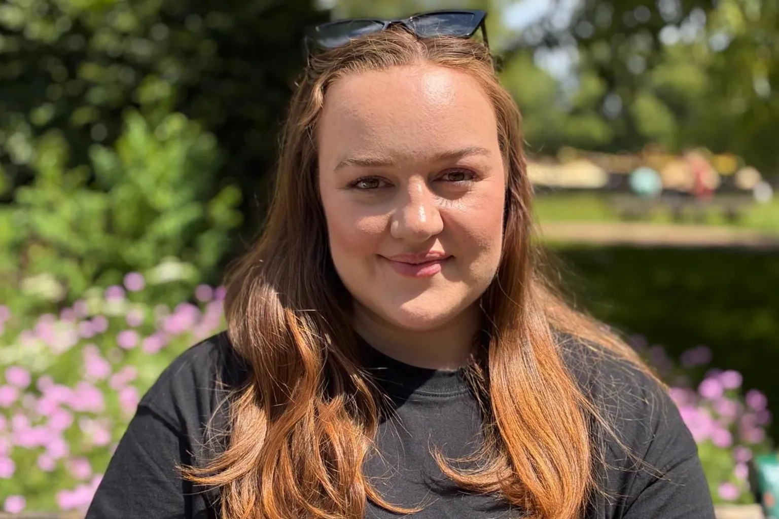 BBC Wales A woman in her late twenties with long hair, wearing a black top, smiling at the camera. She's sat in a public park with greenery behind her.