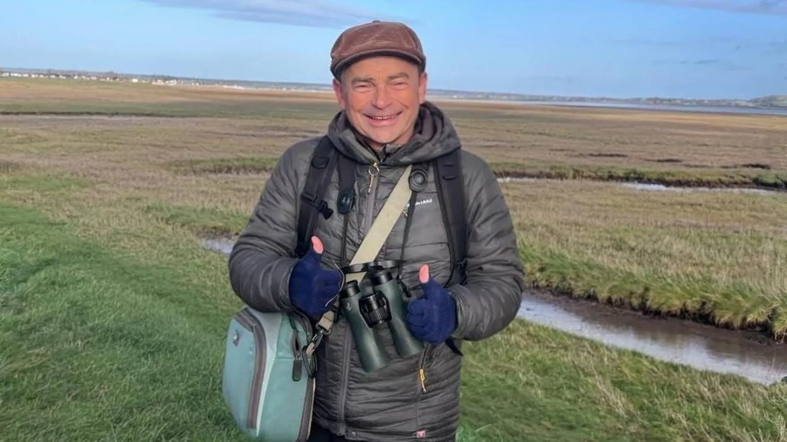 Marc Hughes Simon smiles as he holds binoculars in his hands. He stands in front of a green nature reserve. Simon wears a grey jacket, a blue shoulder bag, navy gloves and a brown cap.