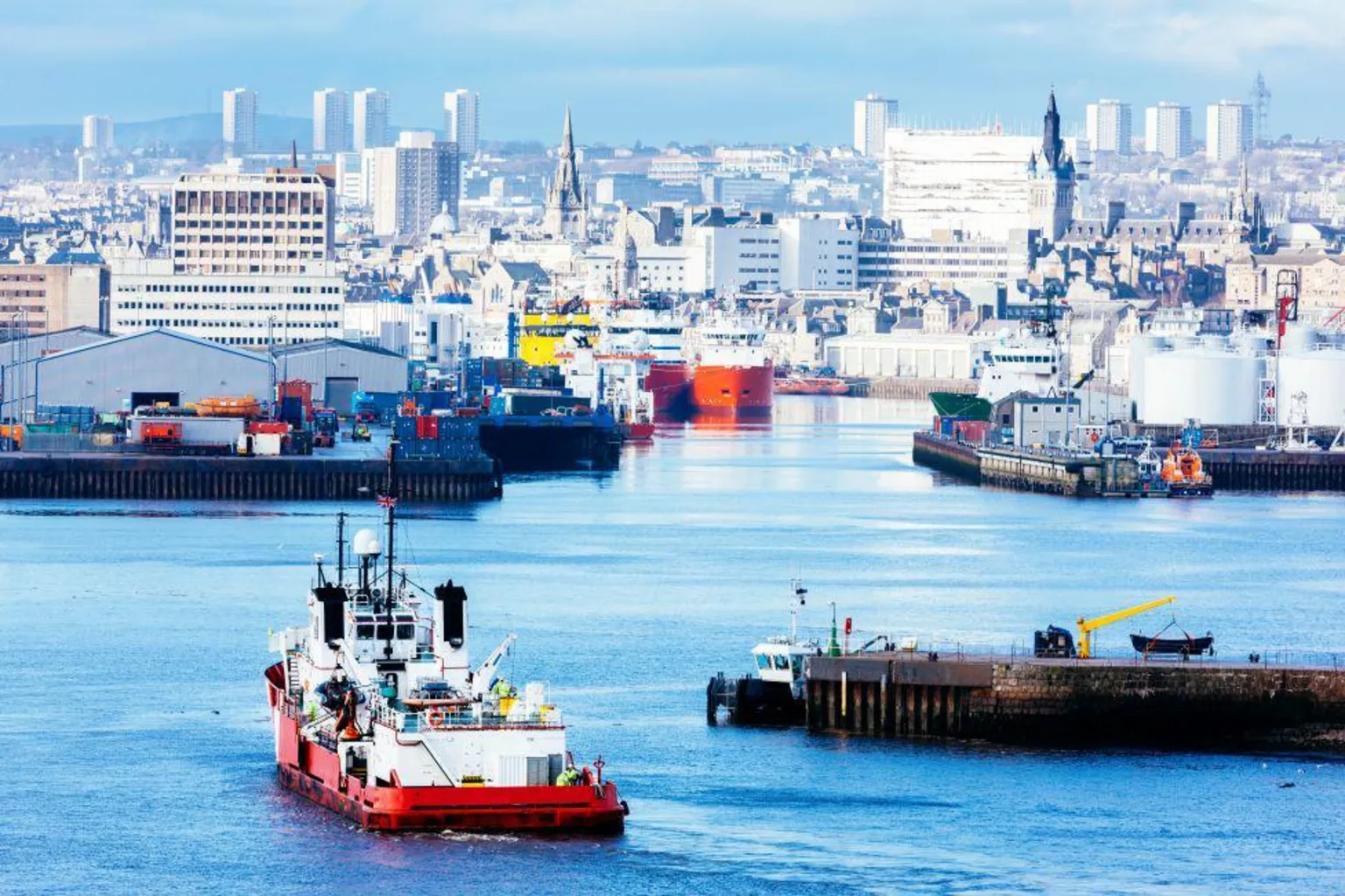 A vessel sailing into Aberdeen Harbour, further boats are in the distance along with the city skyline, including several high-rises.
