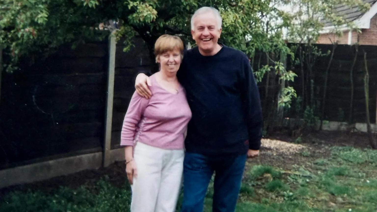 Stephen Hindley Judith and Steve Hindley standing together in a garden, with Judith wearing a light pink top and pale trousers and Steve in a dark jumper and jeans, his arm around Judith, in front of a wooden fence and leafy trees.