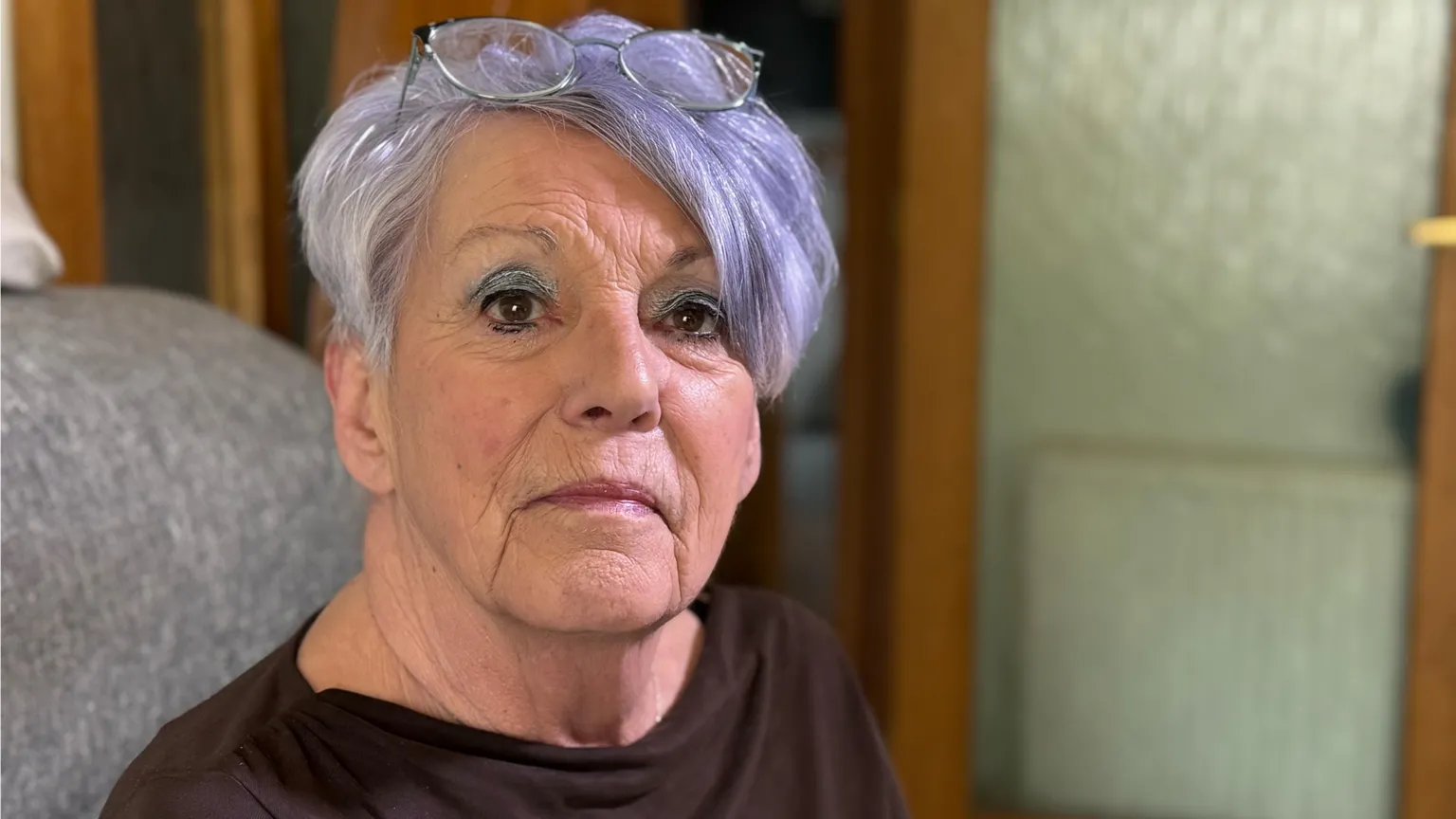 Close-up portrait of a Jan Lawden who has short purple hair and round glasses resting on top of her head. She is sitting indoors and wearing a dark top, in front of a softly blurred background of wooden furniture and a glass-fronted cabinet.
