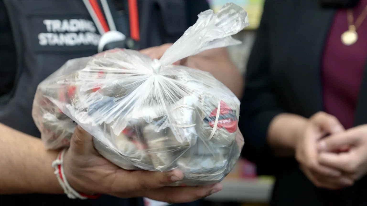 Close-up of a tied, transparent plastic bag filled with empty metal snuff containers being handed between two people. One person wears a dark shirt with partially visible text reading “TRADING STANDARDS,” while another person’s hands and a pendant necklace appear in the background.