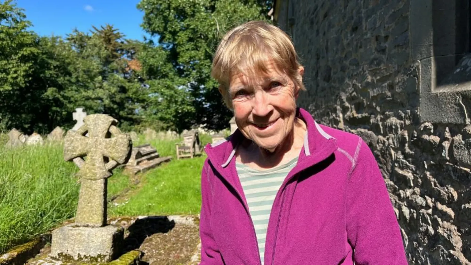 Ellen Knight/BBC Kate pictured in the churchyard. She is wearing a bright pink fleece with a grey striped top underneath. To her right is a Celtic cross gravestone, and to her left is the grey stone wall of the church. The sky is blue and there is strong sunshine.
