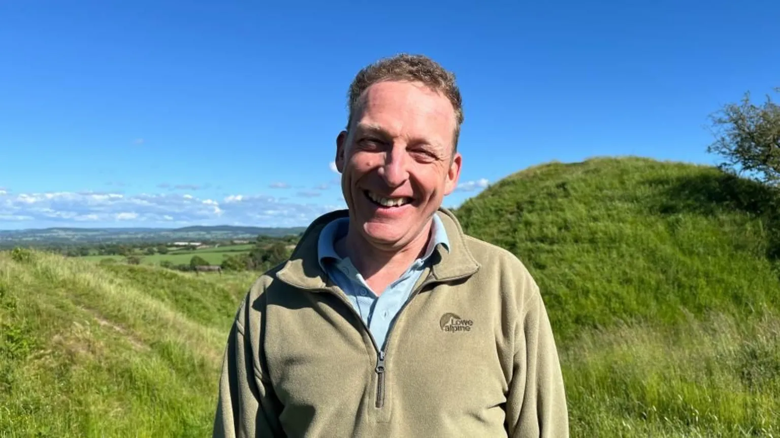 Ellen Knight/BBC Phil pictured looking into the camera and smiling. He is wearing a green-beige fleece with a blue shirt underneath. Behind him is a huge earthen mound, covered in grass, with more slopes leading away from it. The sky is bright blue and there is strong sunshine.