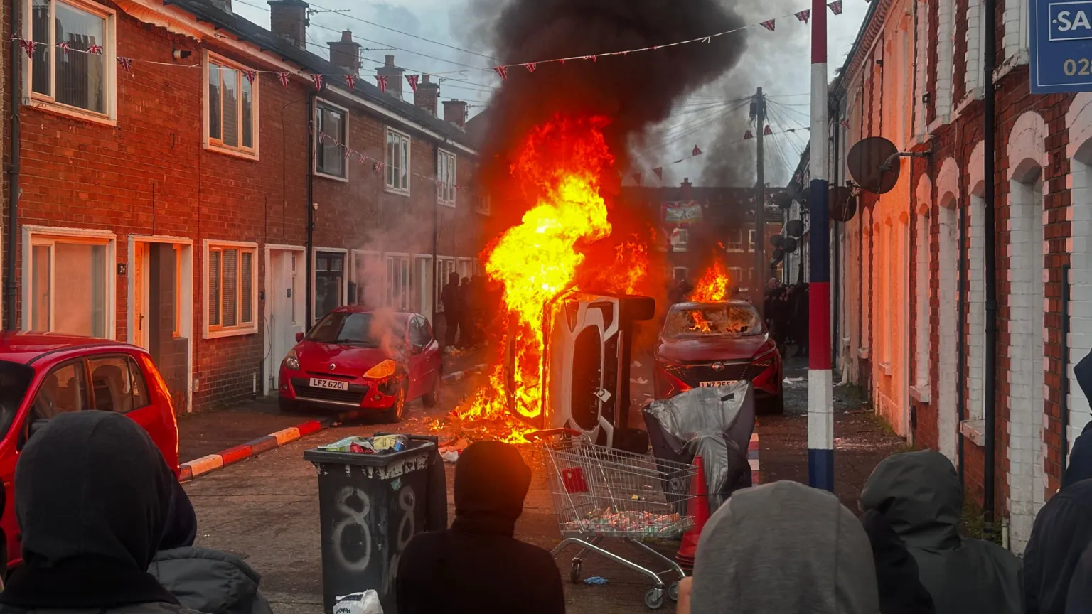 An overturned vehicle burns in the middle of a residential street as thick black smoke rises into the sky. Flames have spread to a nearby car, while a crowd of hooded onlookers watches from behind makeshift debris on the road. Union flags hang overhead between rows of terraced houses.