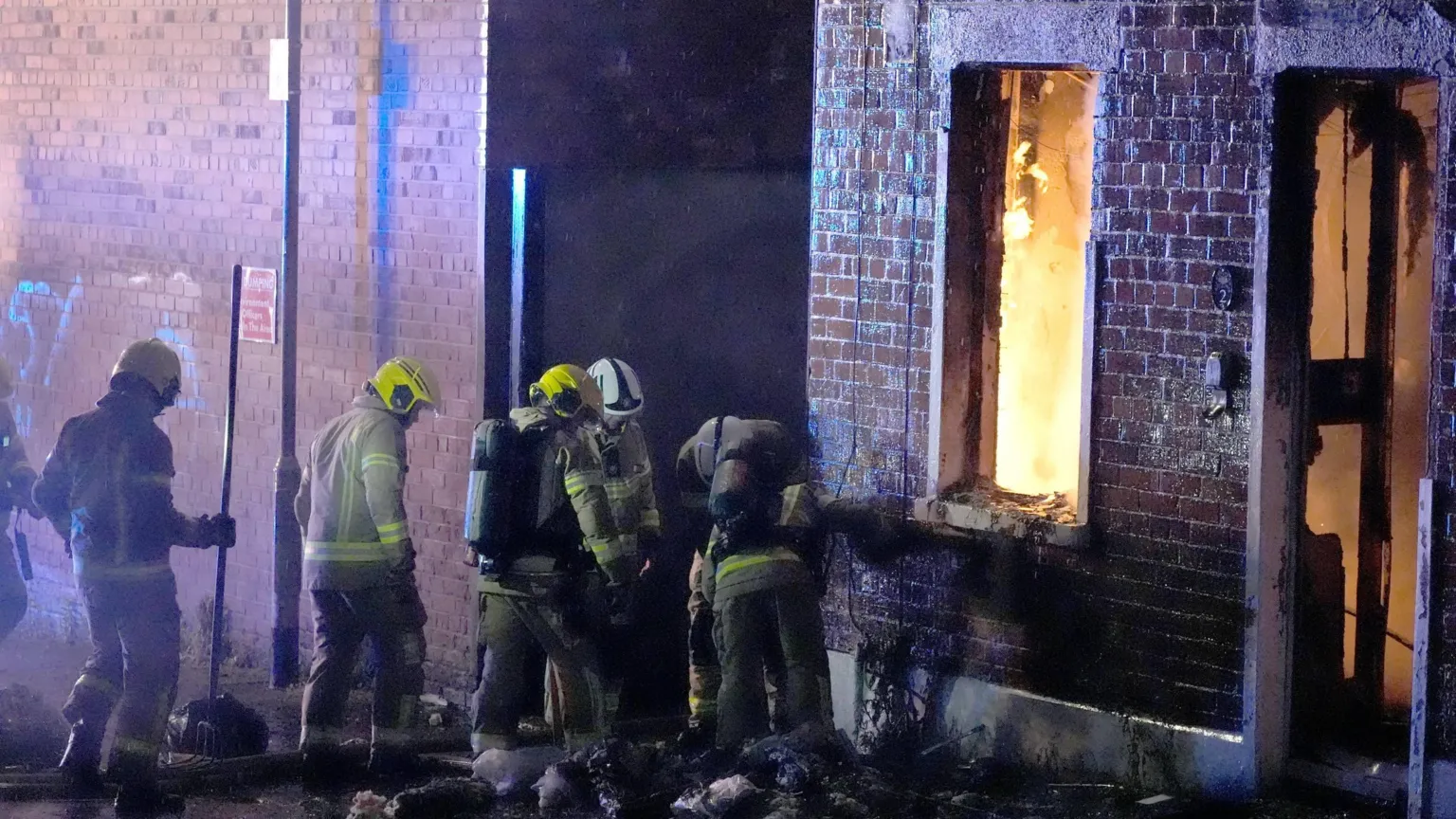 Firefighters work outside a building damaged by fire, with flames visible through a window and doorway. The exterior walls are blackened by smoke as emergency crews respond at the scene during the night.