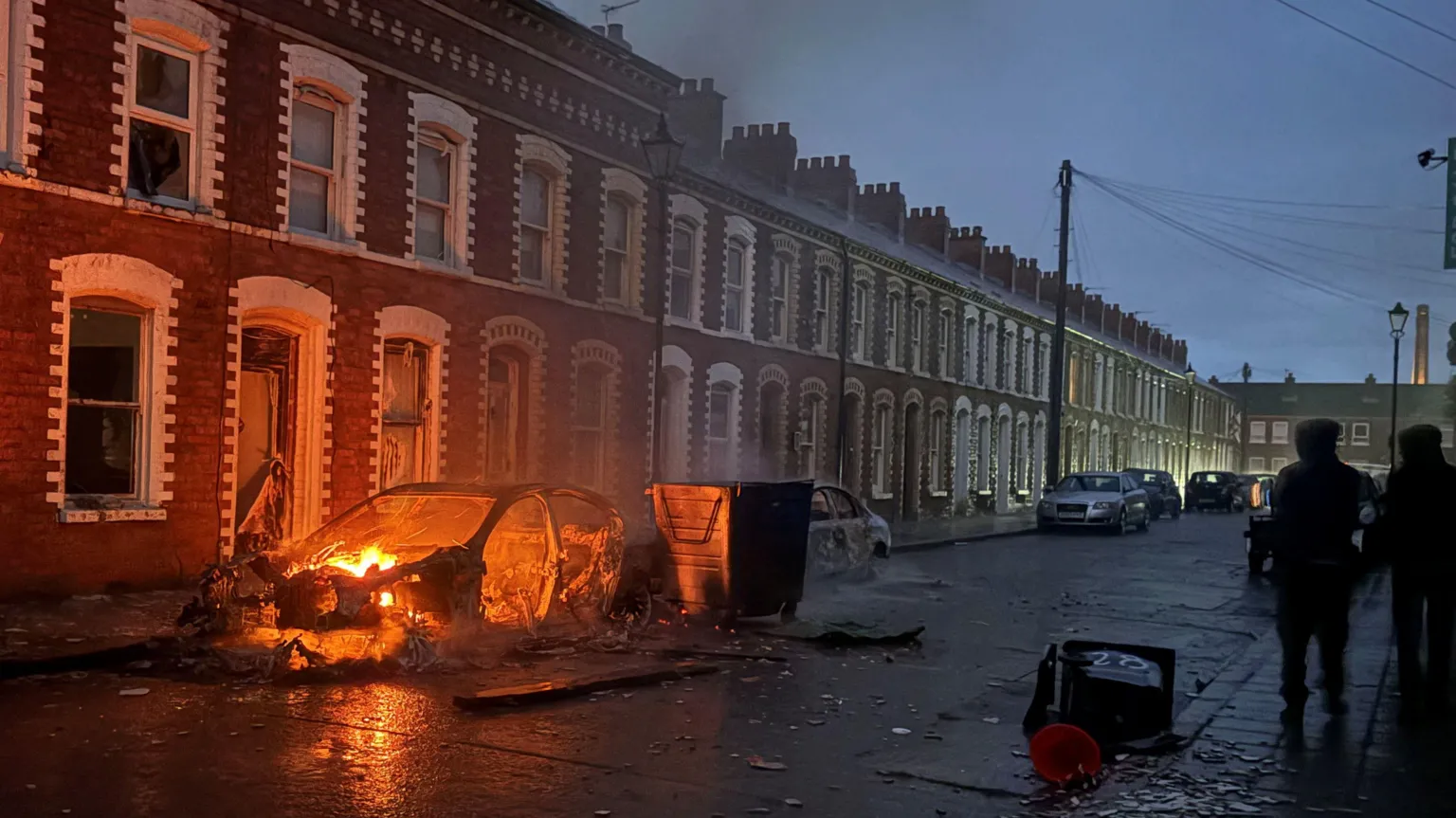 A burned-out car sits outside a row of terraced houses, with smoke still rising from the wreckage. Debris is scattered across the street as a small number of people stand nearby.