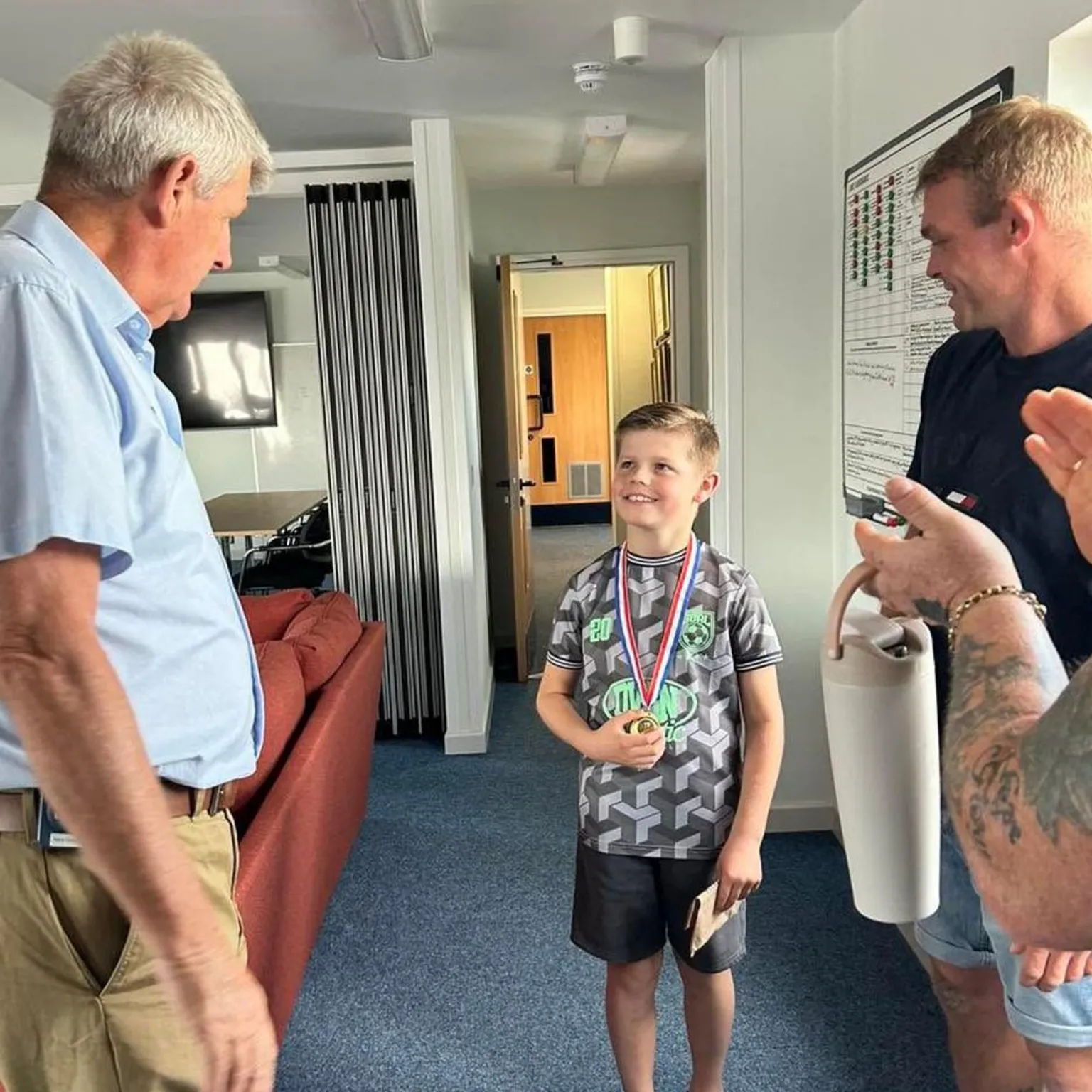 RNLI Barry Dock Marley smiles after being given his medal by an RNLI crew member. His dad and grandad watch on and his grandad claps. 