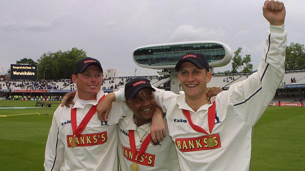 Keith Piper stands in the middle of three players with Ian Bell to his right and Jim Troughton to his left. All three are wearing gold medals on right ribbons around their necks after winning the Benson and Hedges Cup at Lord's. They are all wearing navy blue caps and Troughton has his left arm raised. The media centre at Lord's is visible behind them as is a black digital scoreboard displaying the message
