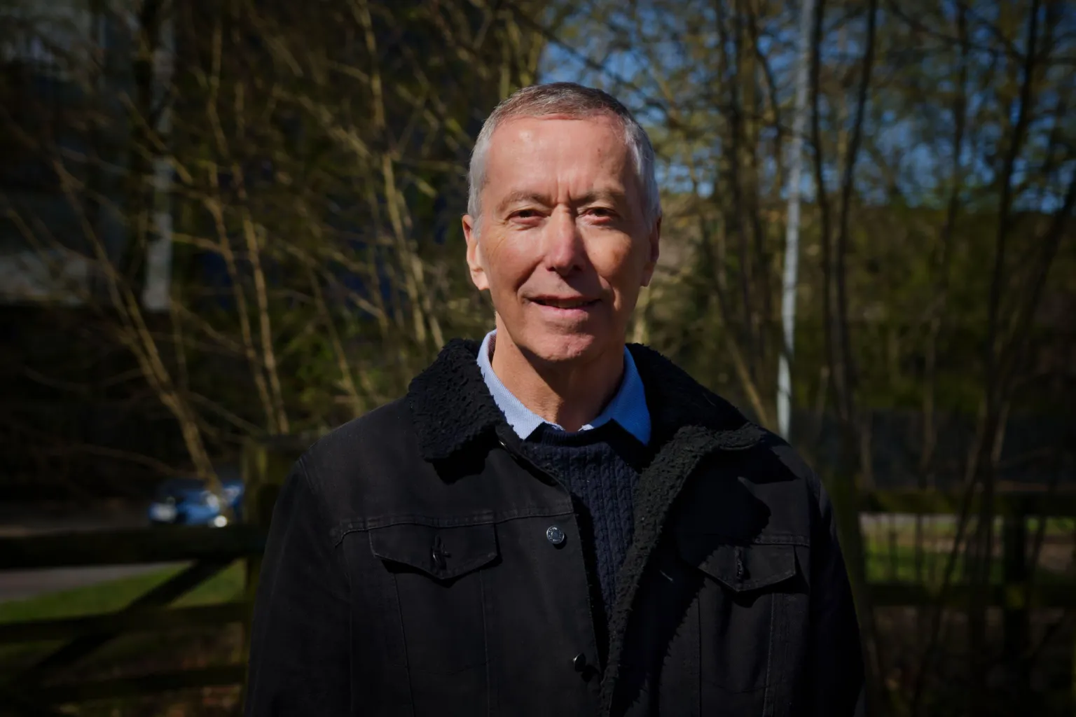 David King has a wry smile towards the camera. He wears a blue Oxford shirt, a blue ribbed woollen jumper and a black denim jacket