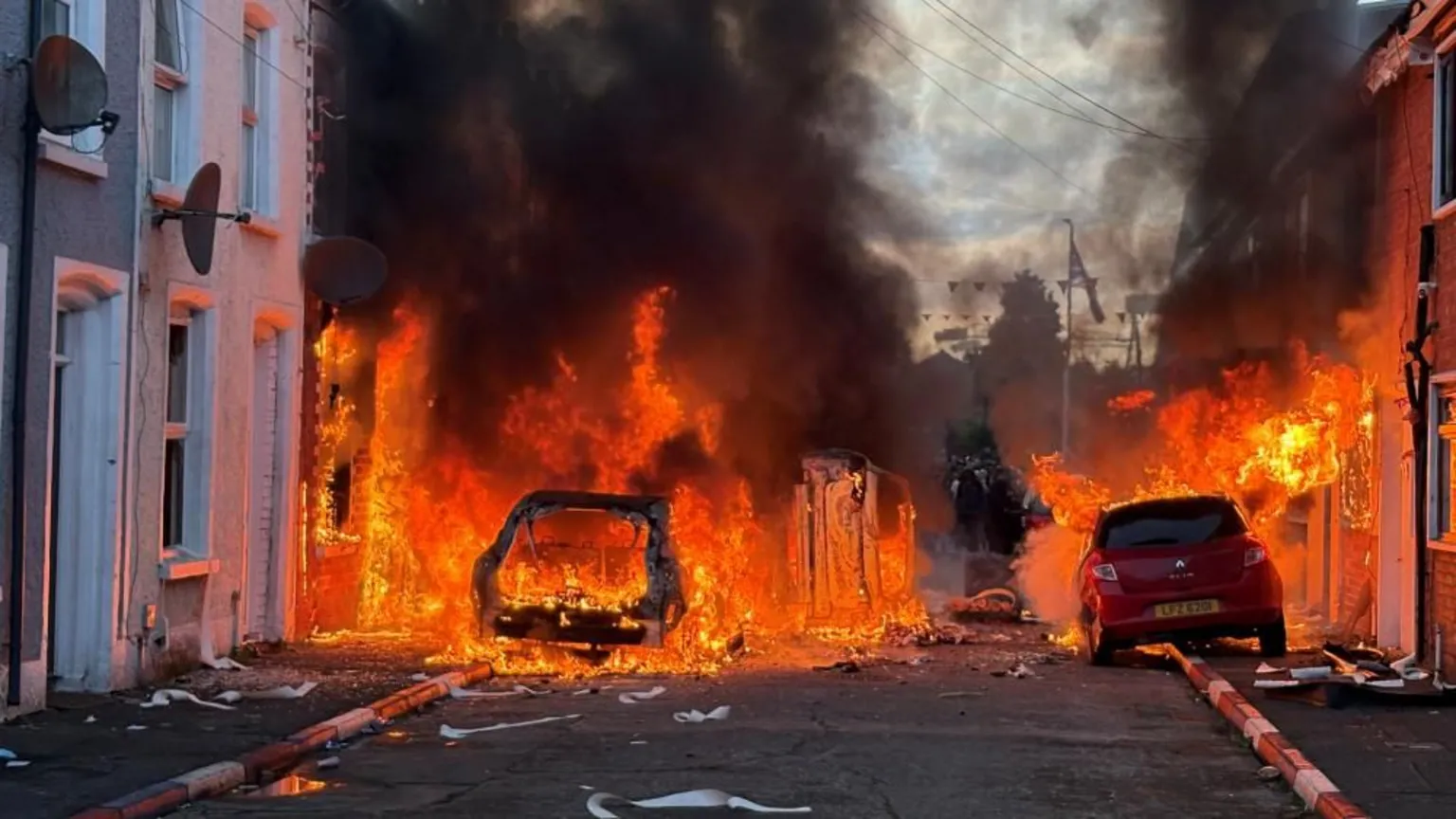 Large orange flames engulf a residential street. Houses and cars are on fire as black smoke billows, obscuring almost everything. One car, on the left, is almost entirely burnt out.