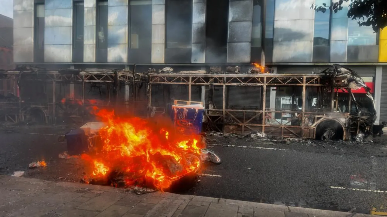 A car in the centre of the photo is fully engulfed in flames, while a shell of a bus - which had been burnt out - sits in the background.