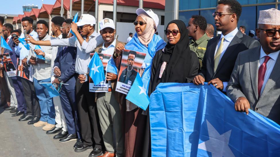 A crowd of people holding Somalia flags (pale blue with a white star) and pictures of Omar Artan