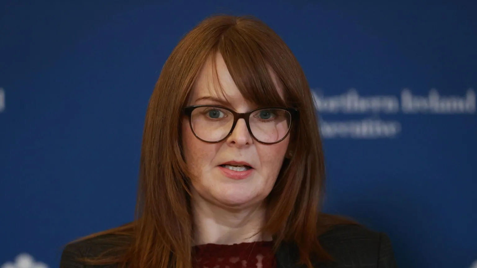 Caoimhe Archibald has auburn hair and glasses standing in front of a blue background