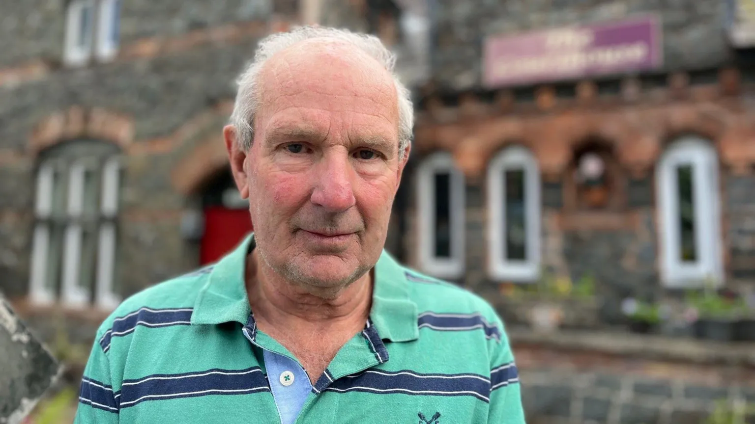 Richard Edwards, looking at the camera. He has thin grey hair, and is wearing a green and blue striped polo shirt. He is stood outside his guesthouse in Betws-y-Coed