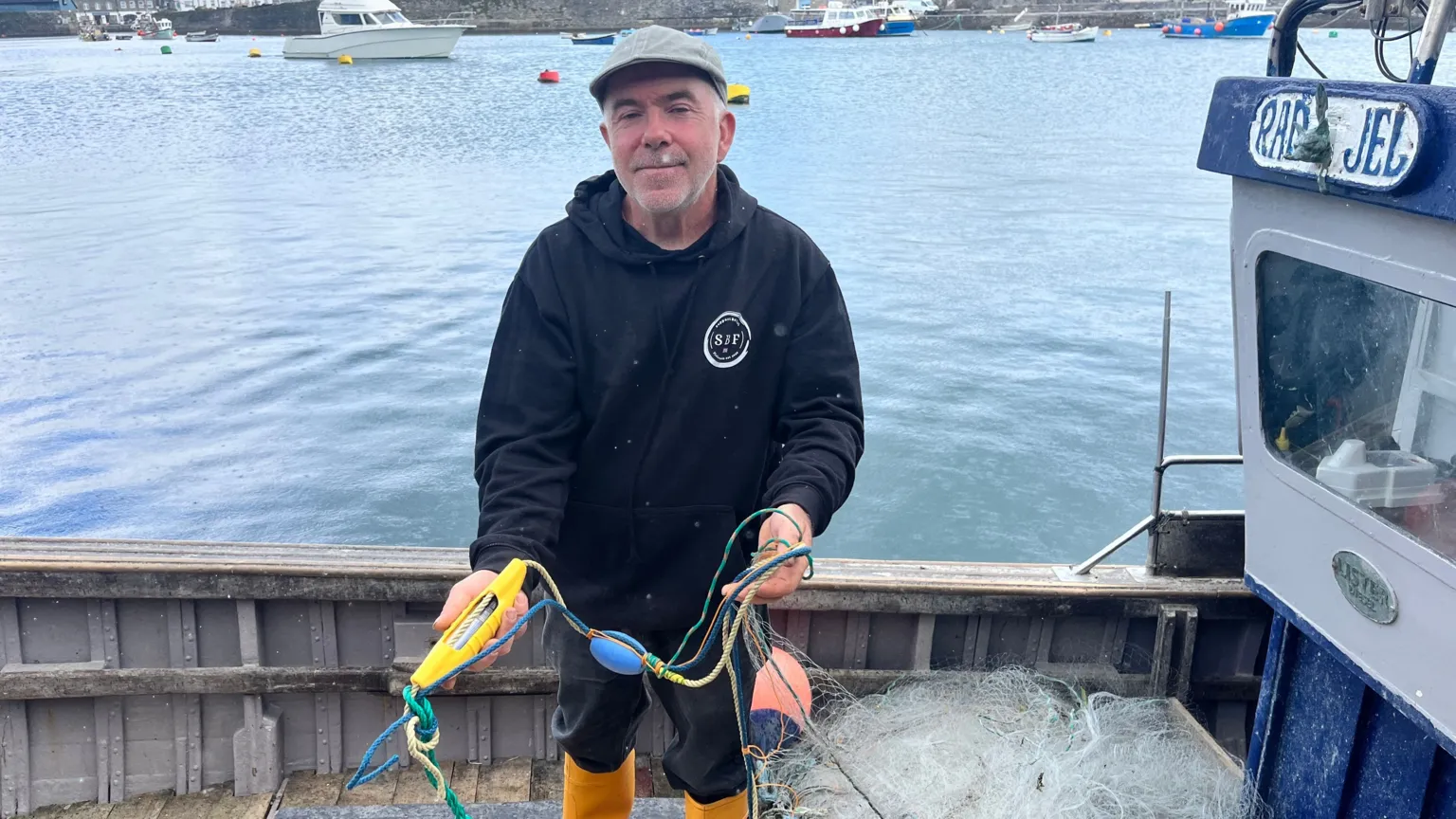Will Shugg stands on his boat holding a pinger, which is yellow and shaped like. a banana, and wearing a flat cap and a black hooded jumper