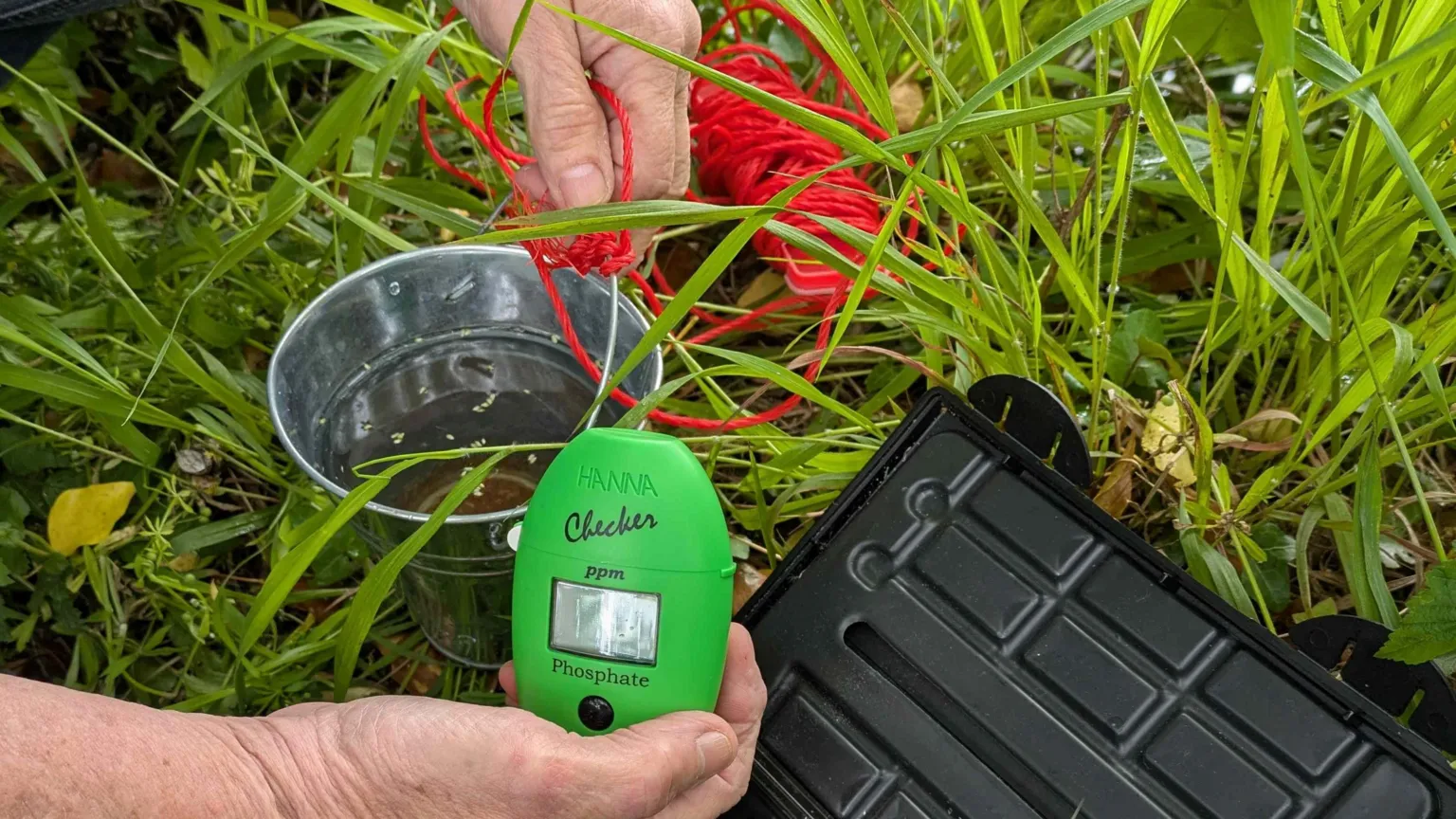 Ruth McDonagh The image shows some of the kit being used to measure different substances in the water. Hands of a man are visible holding a green, handheld, Hanna branded phosphate measuring device. A small silver stainless steel bucket with red string attached to it and a black plastic case for the kit are in the background as well as grass and foliage on the ground.