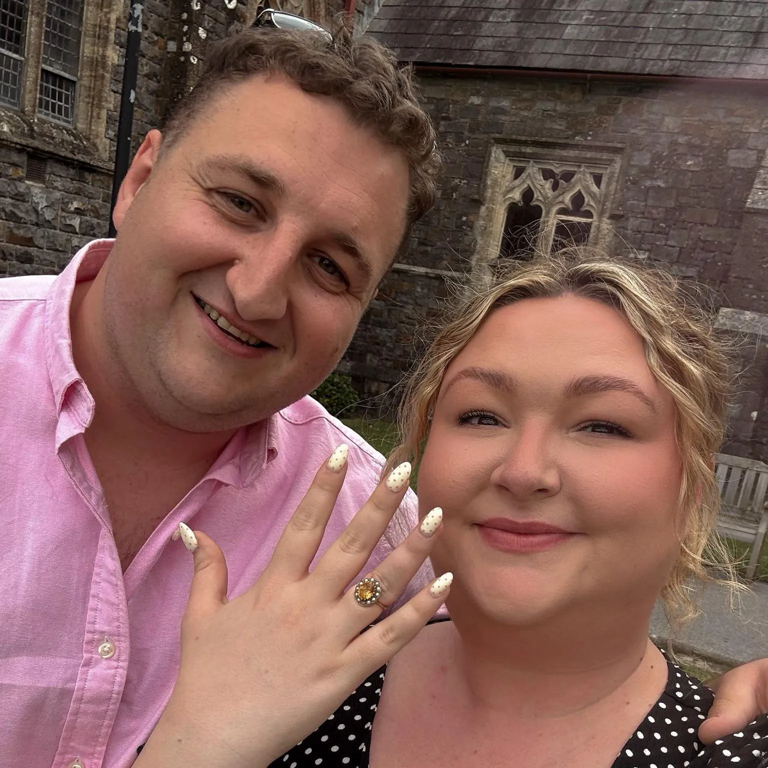 Kayley Stead Kayley showing off her engagement ring with hand up wearing a black dress with white polka dots and Richard with arm over her shoulder in pink shirt