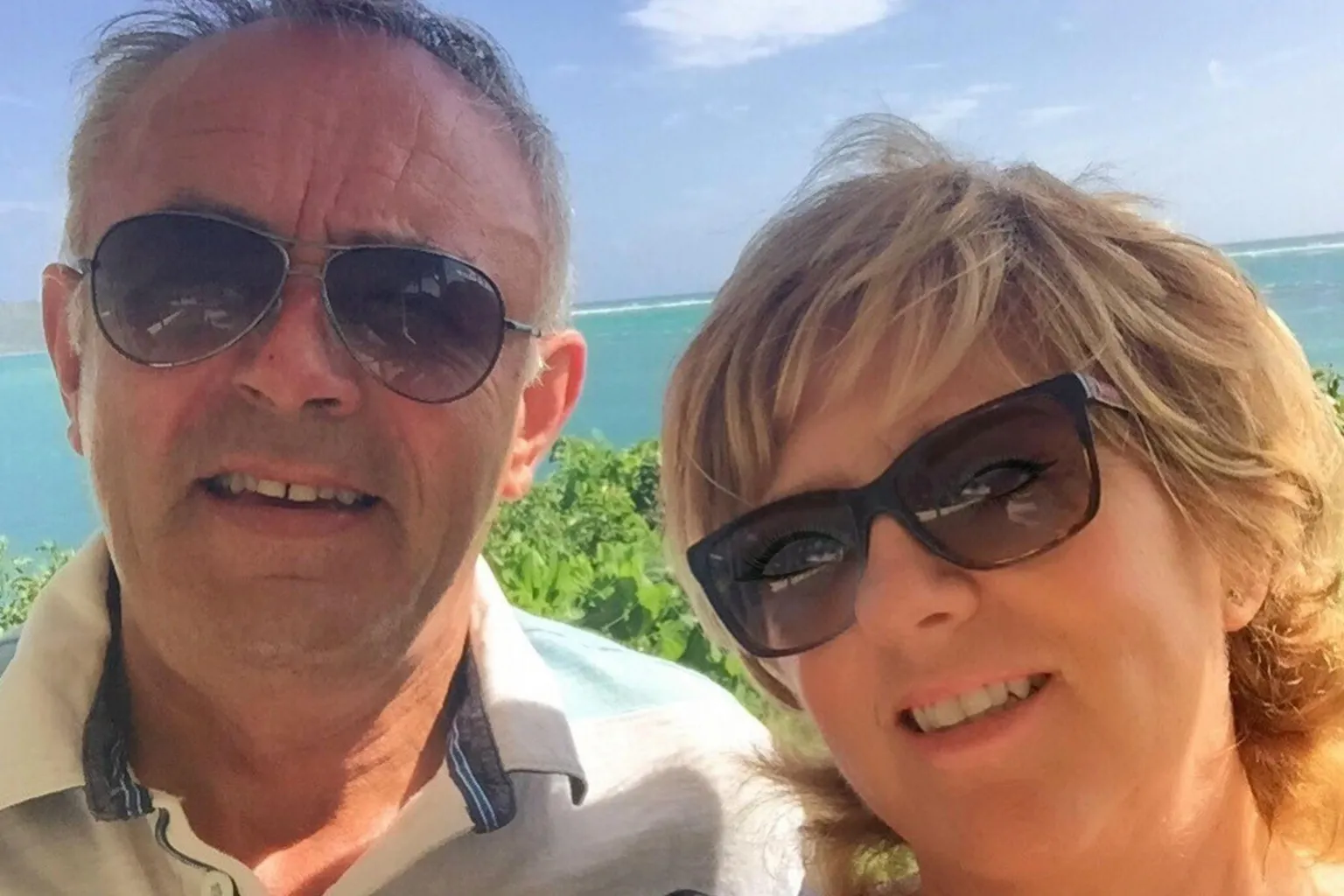 PA Real Life Hugh and his wife Rachel smiling together in sunglasses, with the sea behind them
