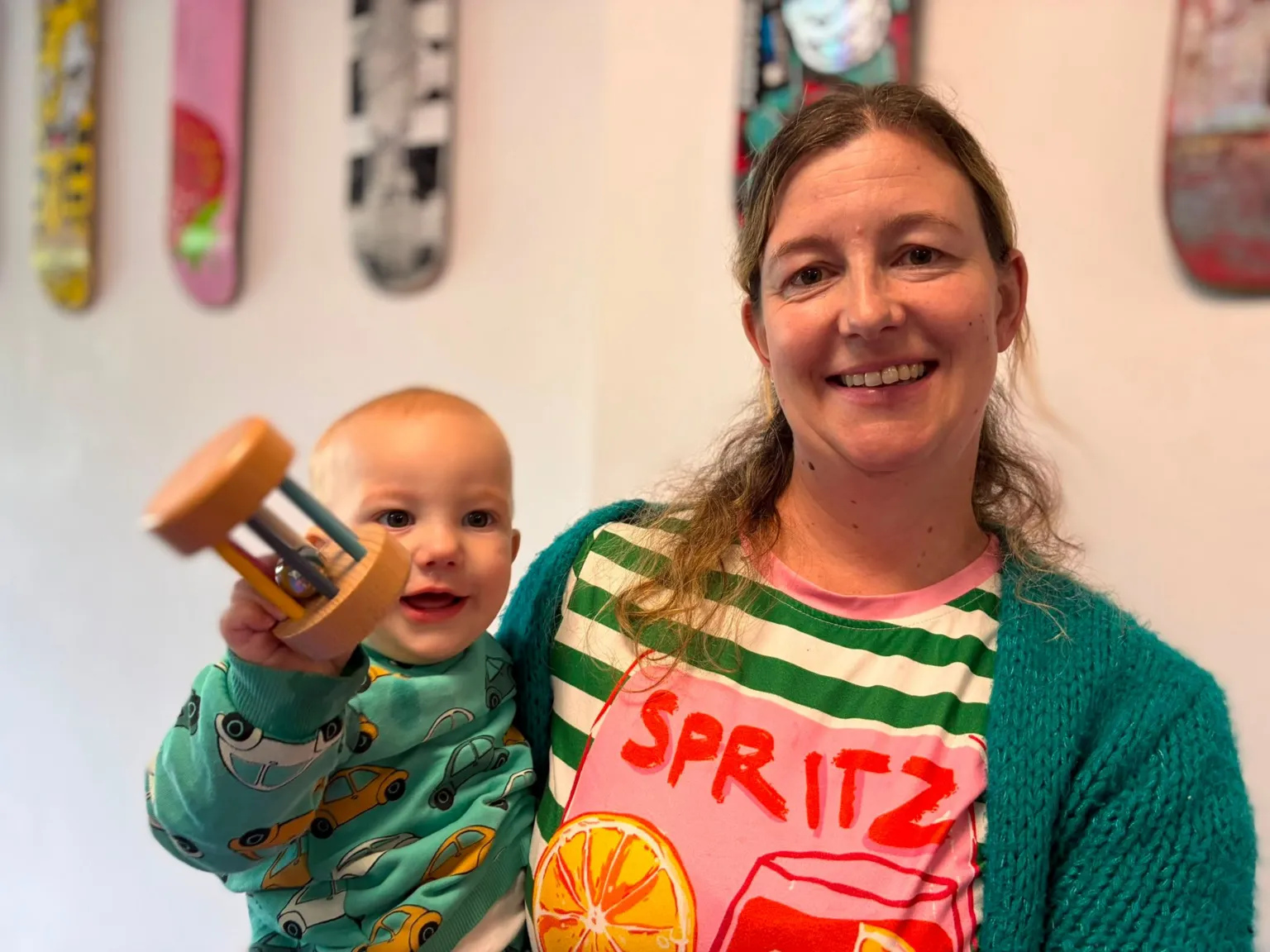 Joshua us on the left in a green jumper with cars on it and is being held by mum, Sarah, in a greed cardigan, green and white striped top with a drawn glass with red liquid and a slice of orange with the word 'Spritz'