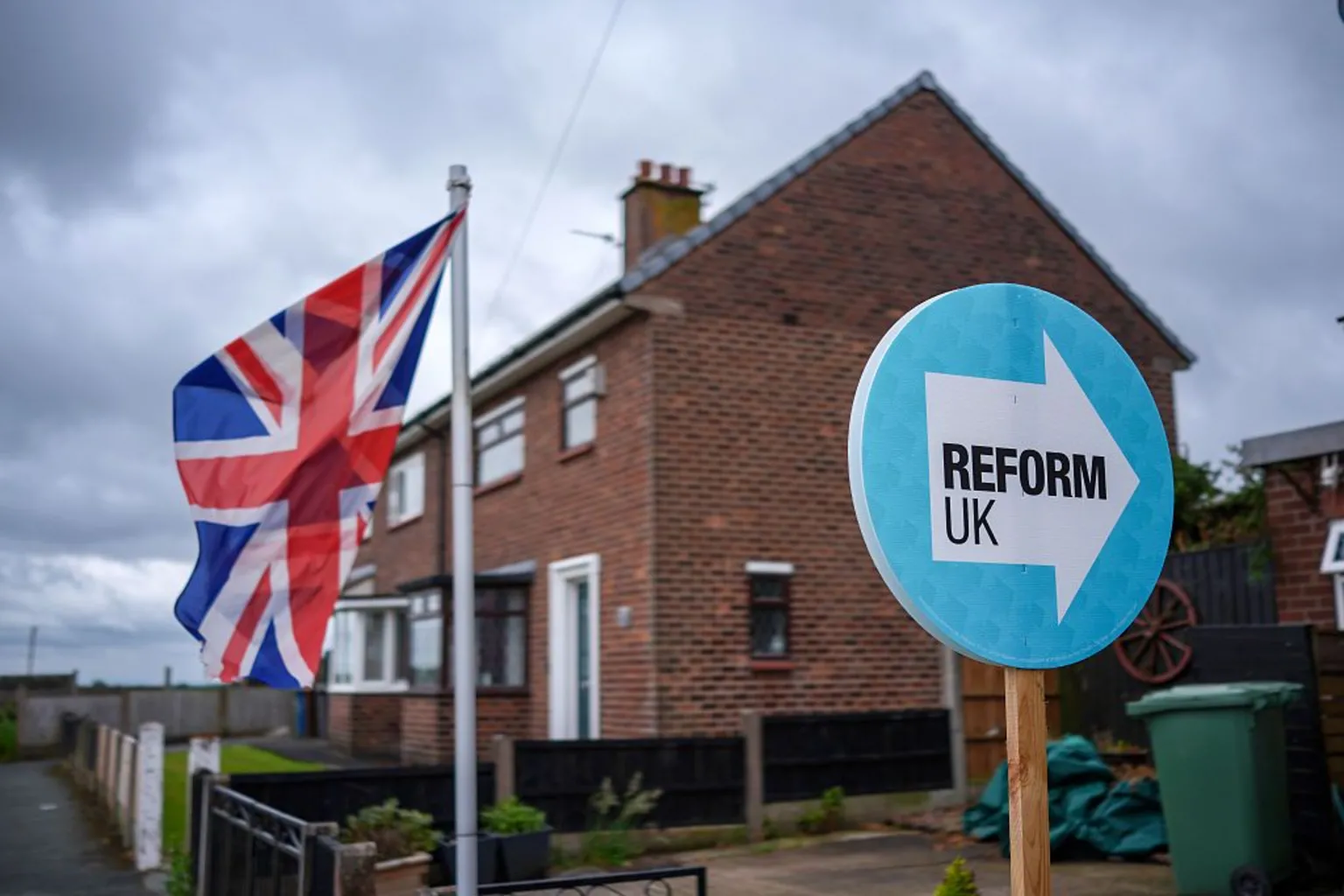  A light blue Reform UK placard next to a Union Jack outside a house.