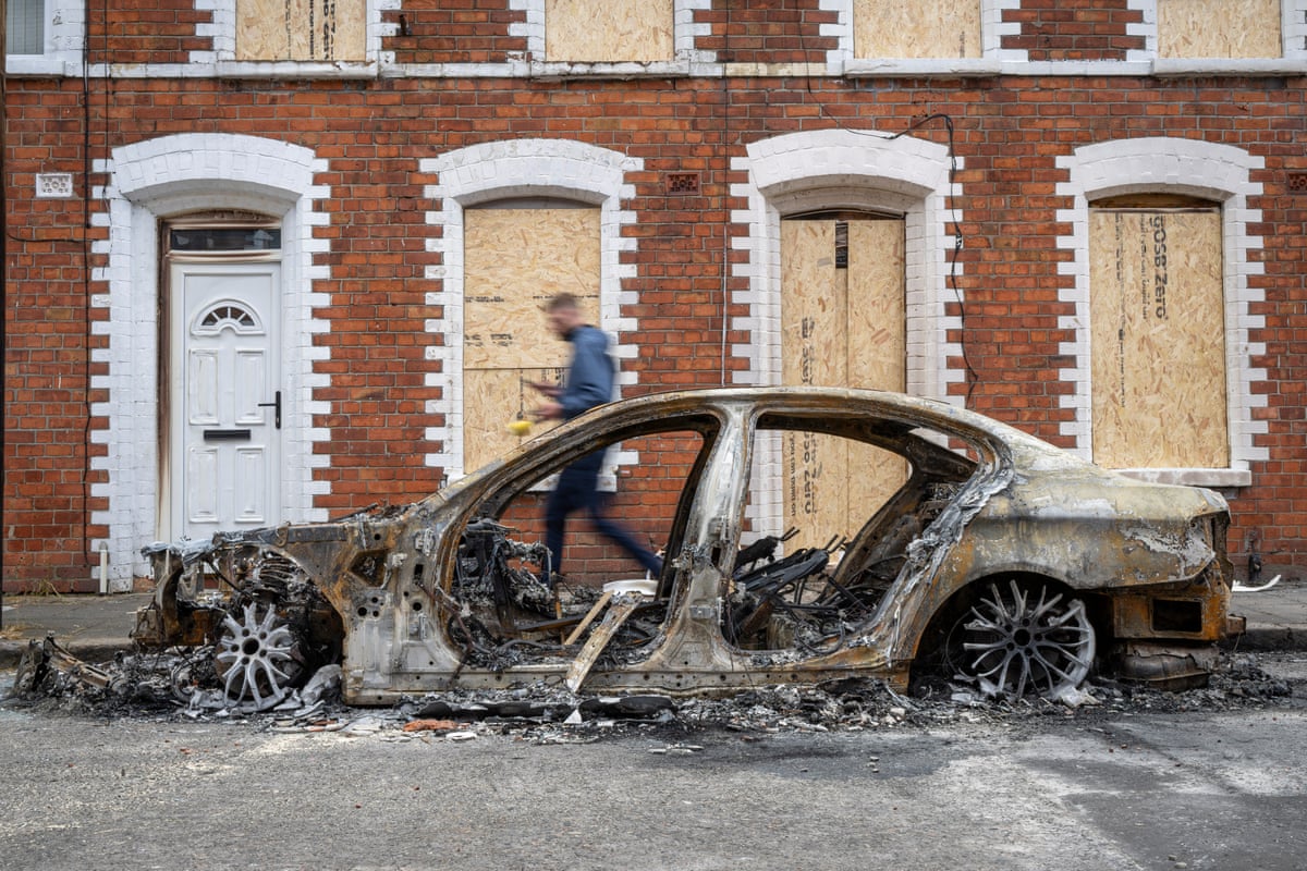 A burnt-out car in front of boarded up houses