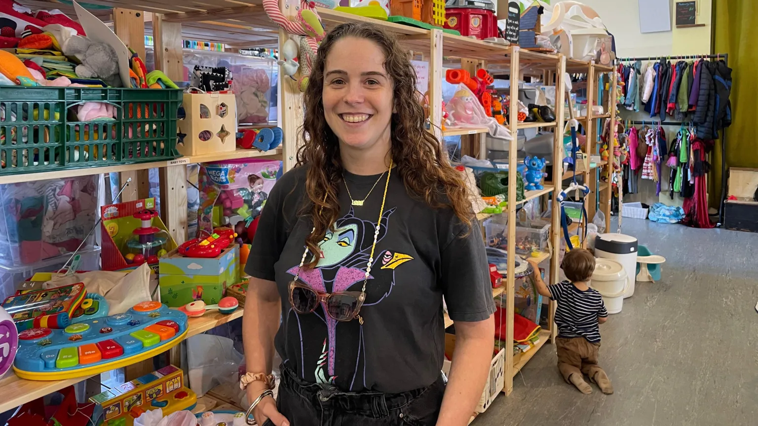 Hannah Jones stands in front of a shelf of toys. She has brown curly hair and is wearing a black tee-shirt featuring Disney character Maleficent.