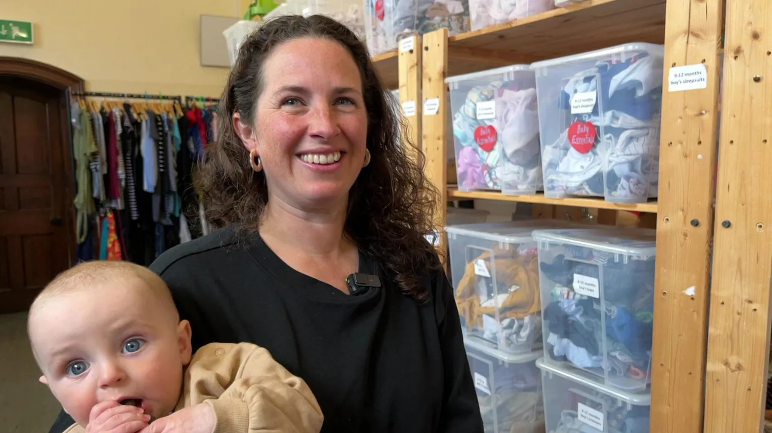 Tabitha Hubbard stands in front of a row of shelves filed with clear plastic boxes. She is holding her seven-month-old son Wilfred. She has brown curly hair and wears a black top.