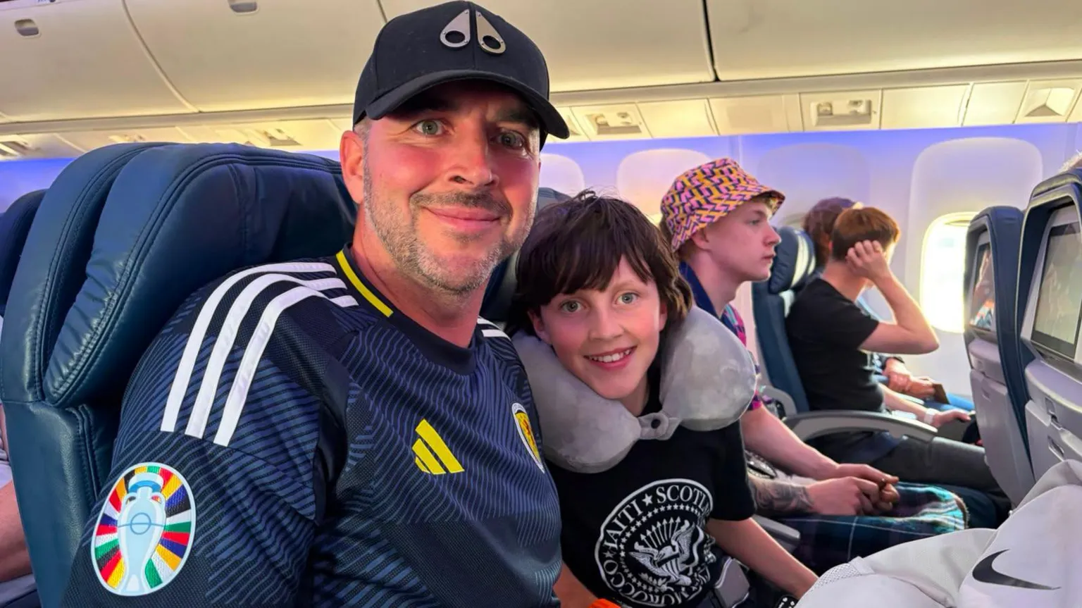 a man wearing a cap and Scotland strip smiling, next to him is a young boy with long brown hair. They are on a plane.