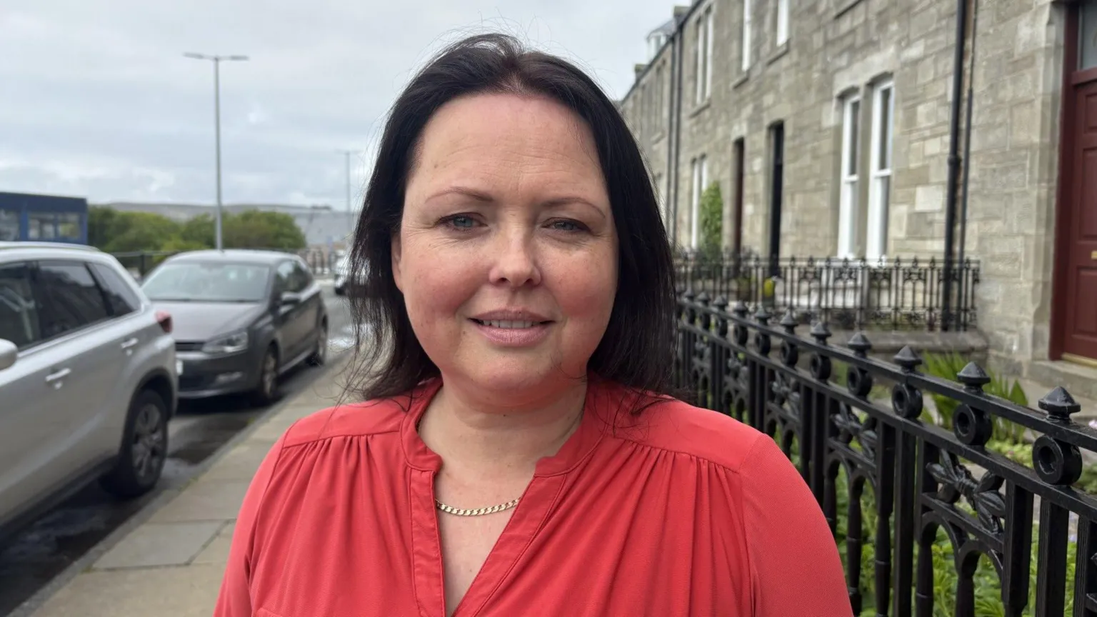 A white woman with black hair. She wearing a red top and a silver necklace, and smiling. The woman is standing on a pavement with a row of houses on one side and parked cars on the other. A cloudy sky. 