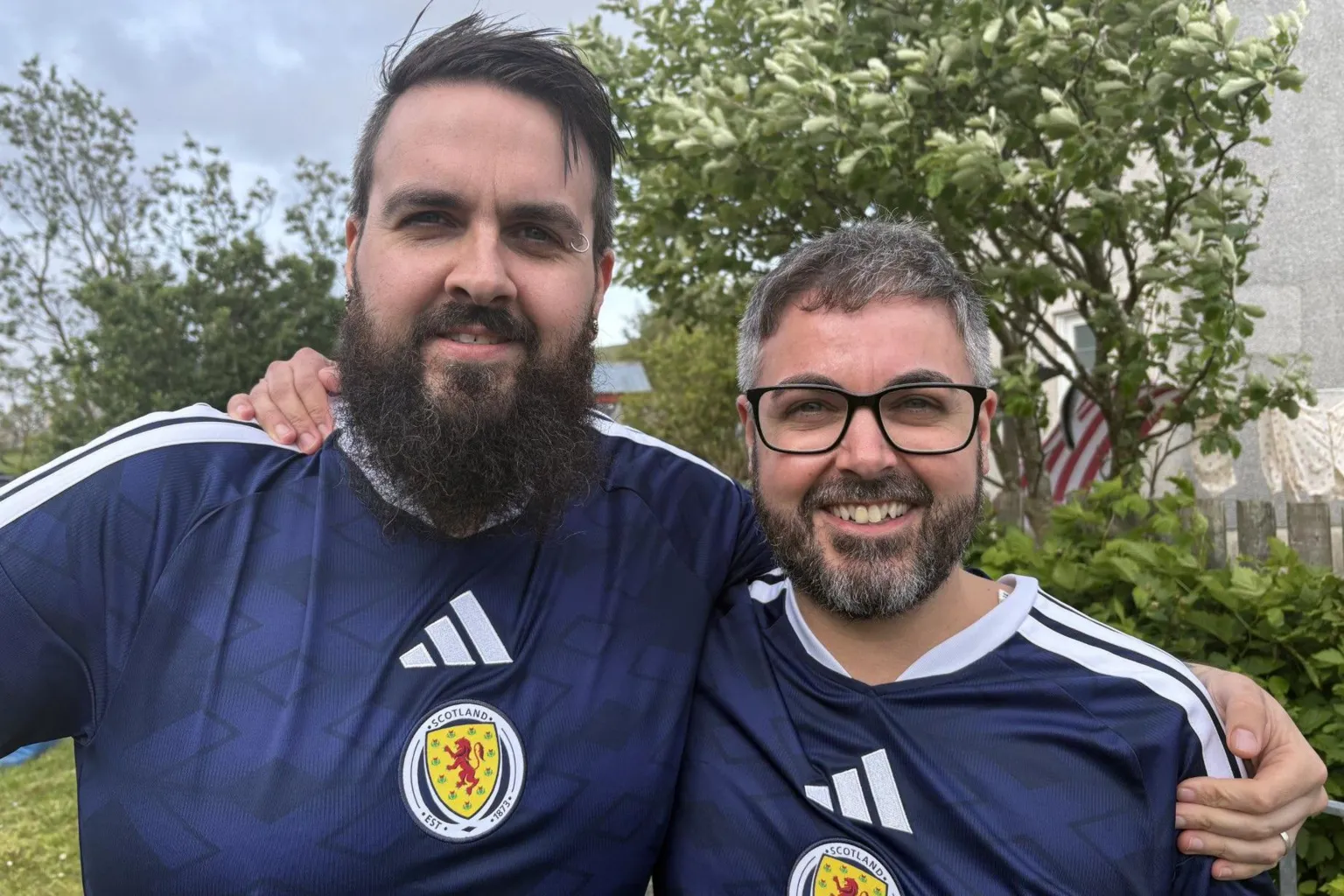 Two men with in blue Scotland shirts with their arms round each other's shoulders. Both men have brown hair and beards, and the man on the right has glasses.