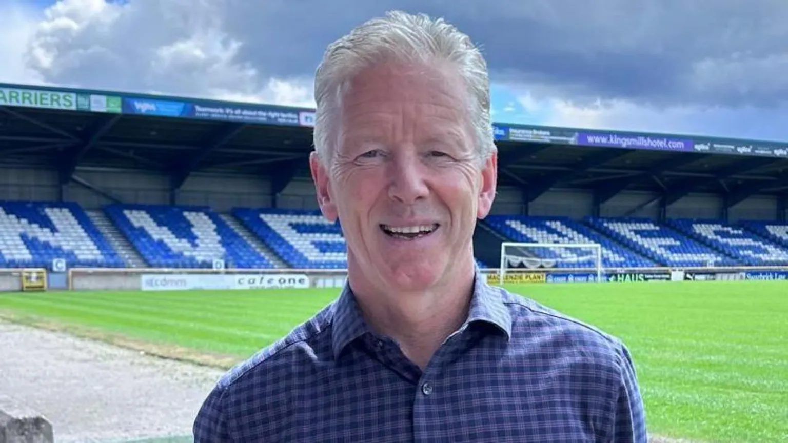 Charlie Christie standing in the Inverness Caley Thistle stadium, with the pitch and a stand visible behind him. He has short grey hair and is wearing a blue checked shirt.