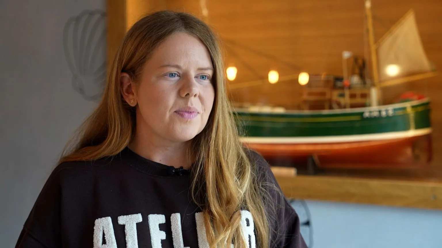 Jo McKenzie, a young woman with long golden hair and a blue and white top. There is a model ship behind her.
