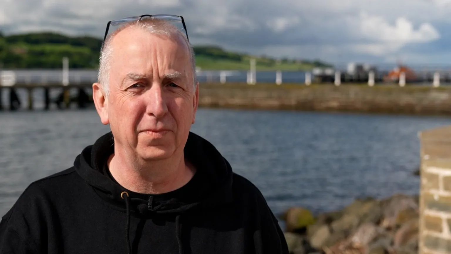 John Kerr stands outside with the Broughty Ferry lifeboat visible in the background. John has short white hair, glasses rested on his head, and is wearing a dark hooded top