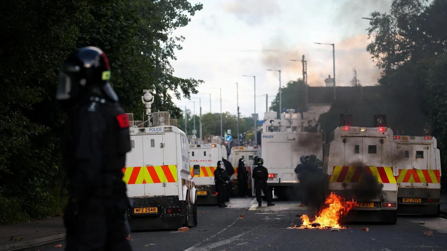 Police officers in dark clothing with black helmets stand beside white land rovers near Sandyknowes roundabout in Newtownabbey. There are flames on the road and black smoke.