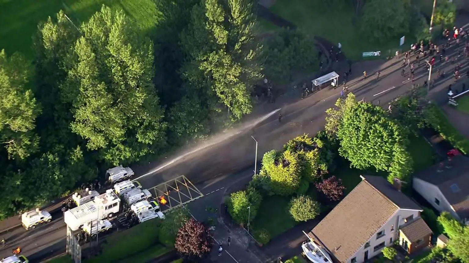 Overhead image of white vehicle squirting water beside white police landrovers near the Sandyknowes roundabout in Newtownabbey.