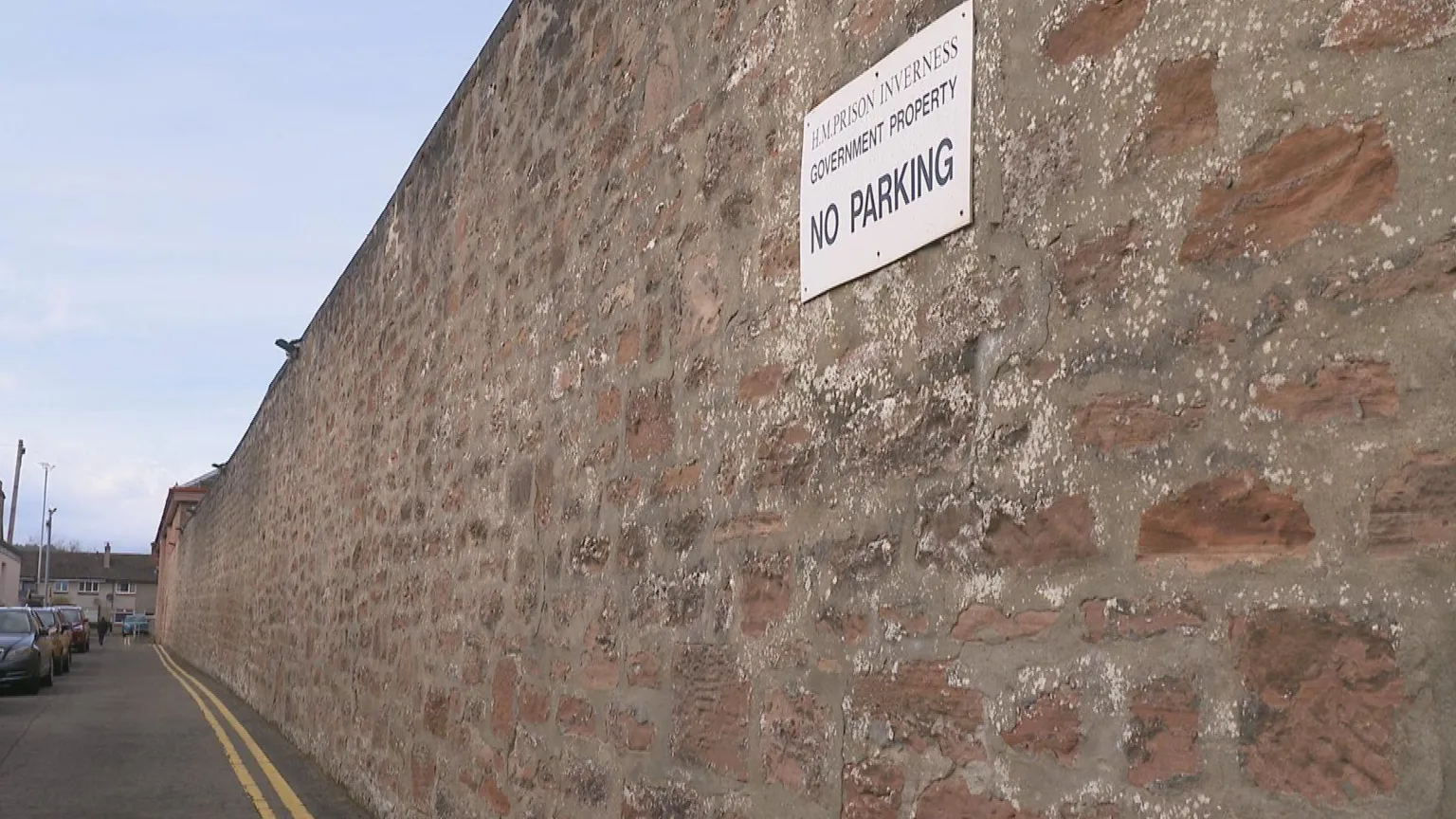 The high red sandstone boundary wall stretches down a narrow street. There is a sign on the wall that has the warning 