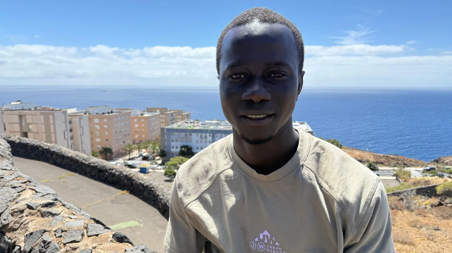 A young Gambian man smiles to the camera in a t-shirt. Behind him is the coastline and apartment buildings