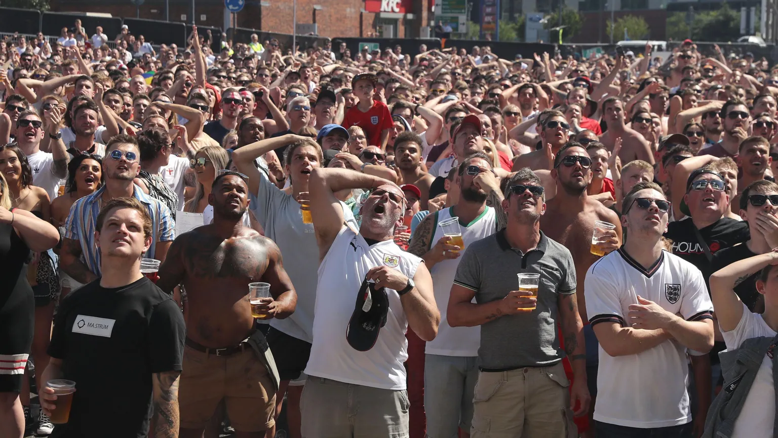 Hundreds of England football supporters stand looking at a big screen as England play. Many of them have their hands on their heads as though a big chance has just been missed. It is a sunny day and some fans have their tops off