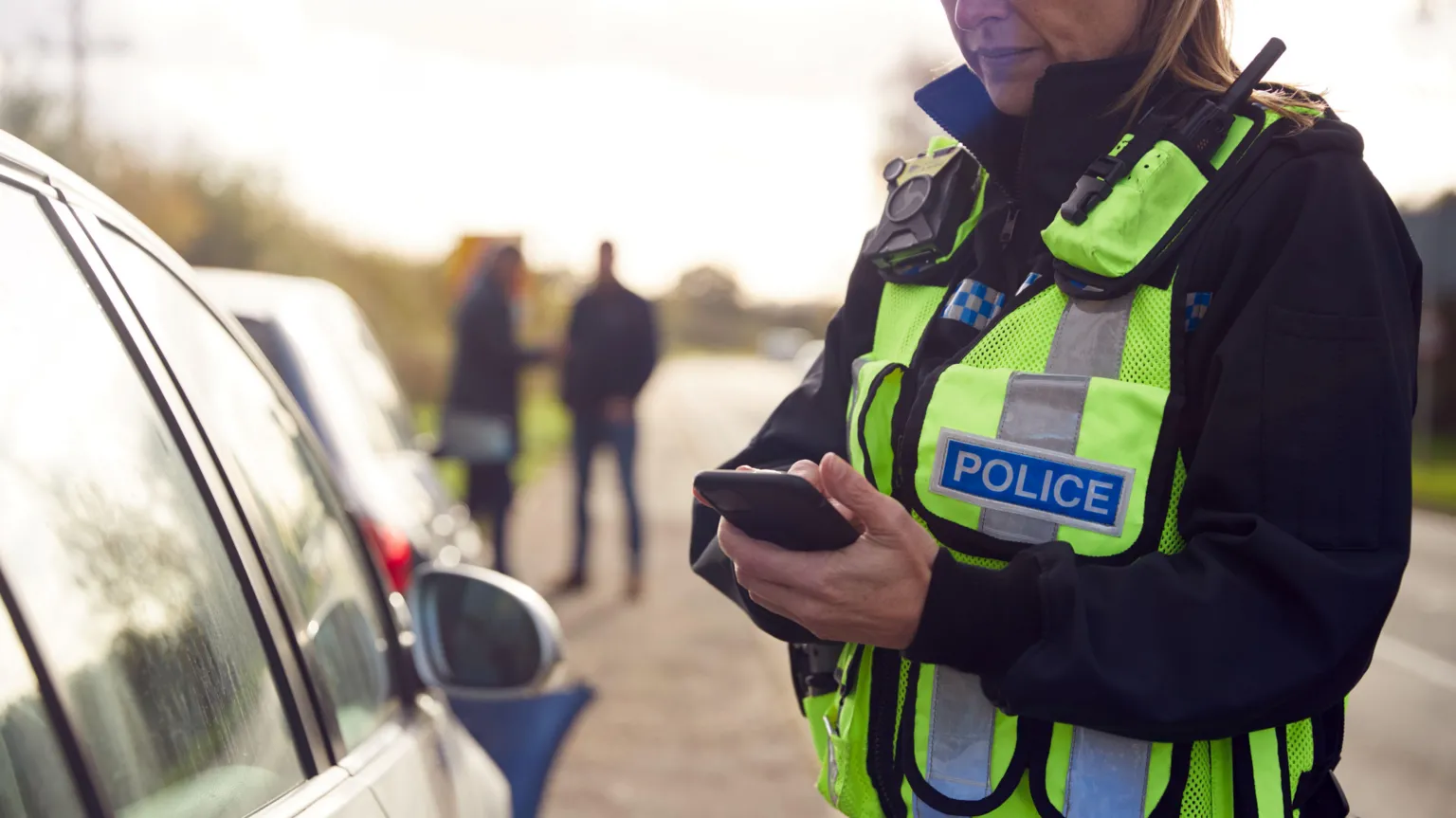 A female police officer in a dark uniform with a yellow high-viz jacket over it stands next to a parked car on a road. She is making notes on her mobile phone
