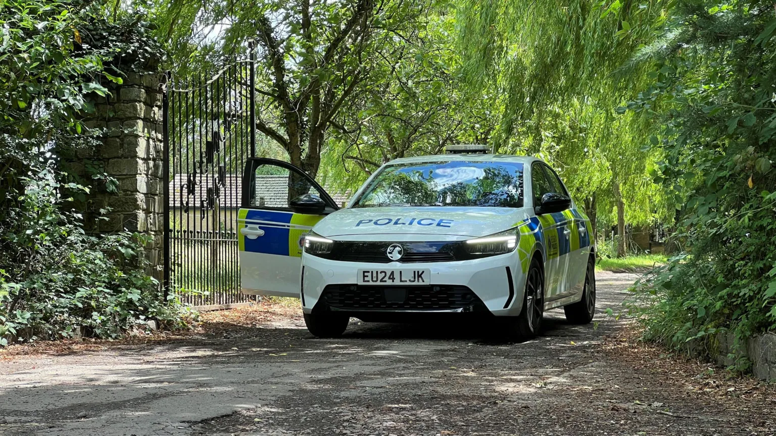 Stuart Woodward/BBC A police car parked on a narrow country track with black iron gates open to the left. The area is leafy and the track is flanked by trees.