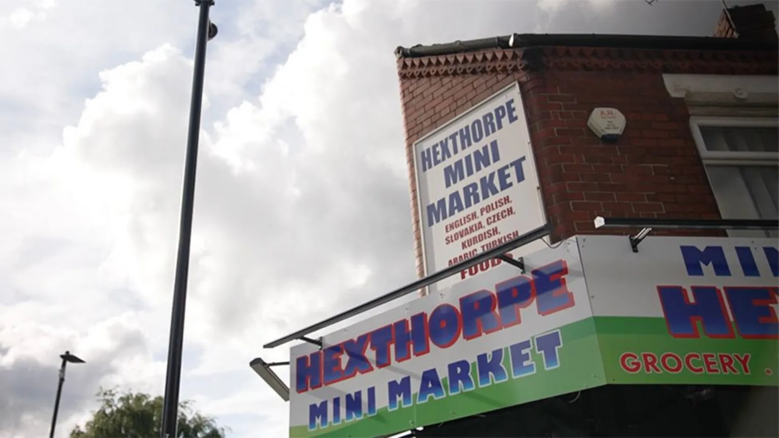 A corner view of a red brick building with a sign reading “Hexthorpe Mini Market,” mounted above a storefront awning. A second sign higher up lists languages including English, Polish, Slovakian, Czech, Kurdish, Arabic, and Turkish. The sky is cloudy, and a tall streetlight stands in the foreground.