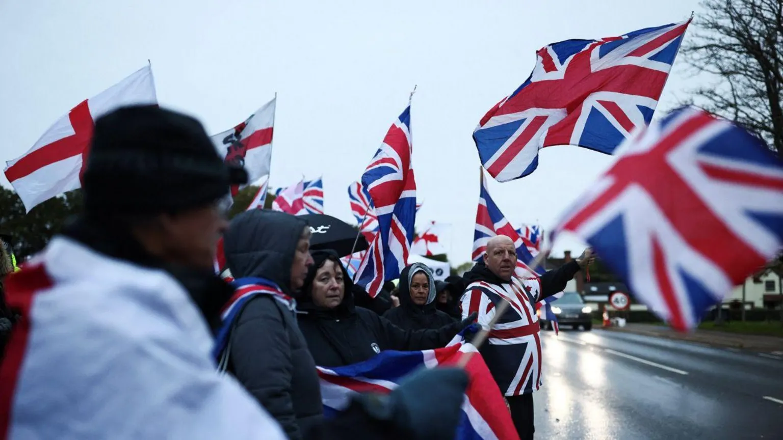 People waving Union Jacks and St George's flags by the side of the road in wet conditions. Some people have hoods and umbrellas up.
