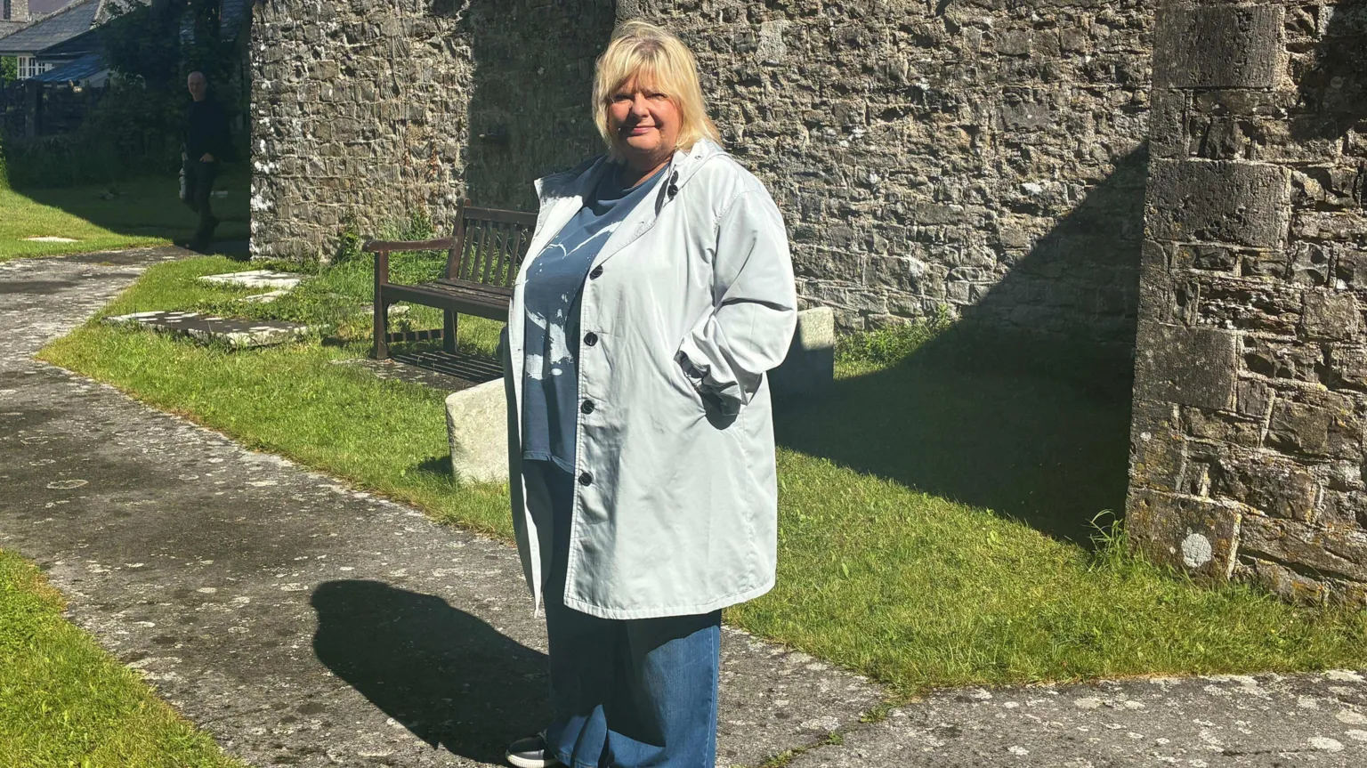 Kieran Molloy A woman with a blonde bob looking at the camera. She has a pale blue raincoat with a blue t-shirt and blue jeans. She is stood in front of a stone building and there is a stone path with grass on either side.