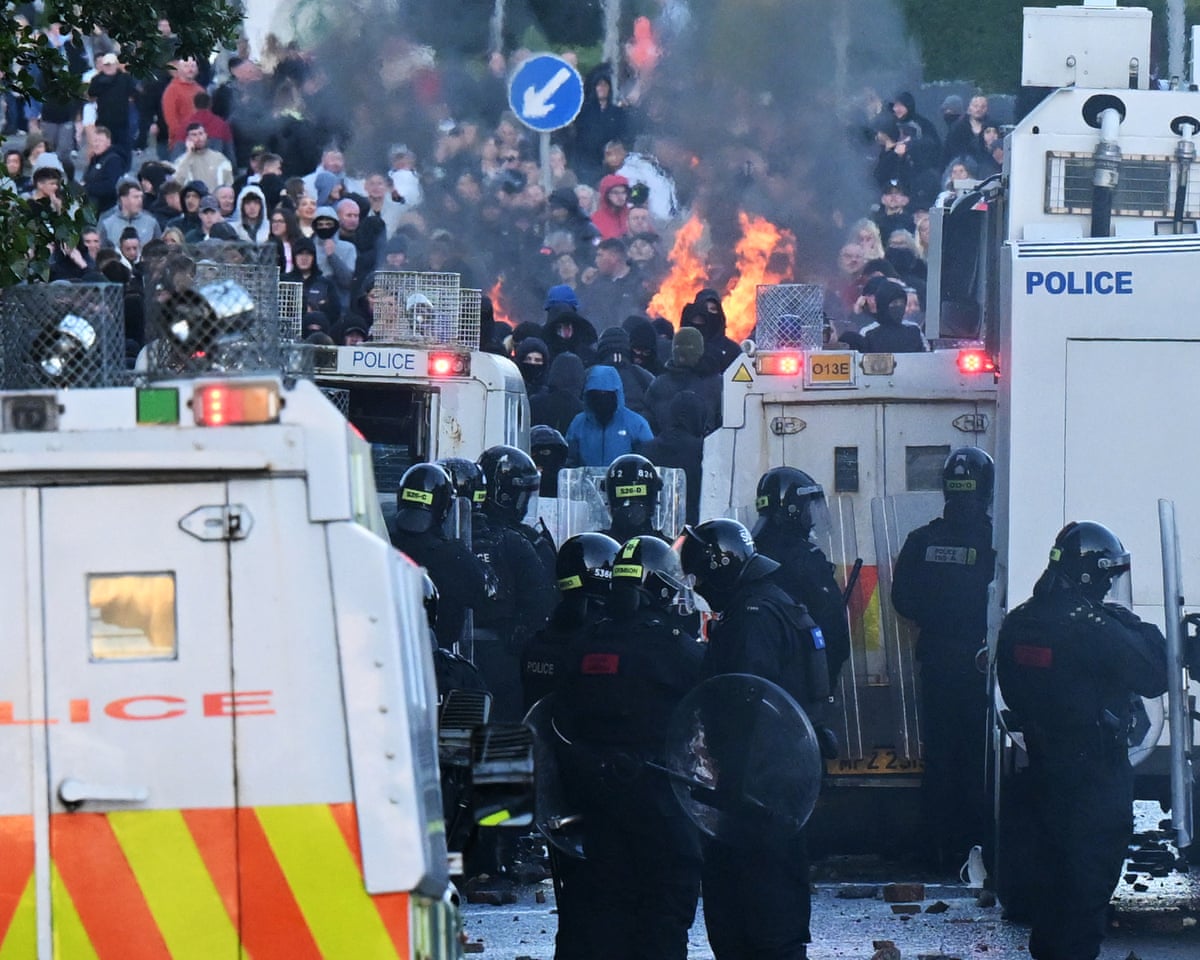 Police stand guard next to vehicles, with protesters and fires in the distance