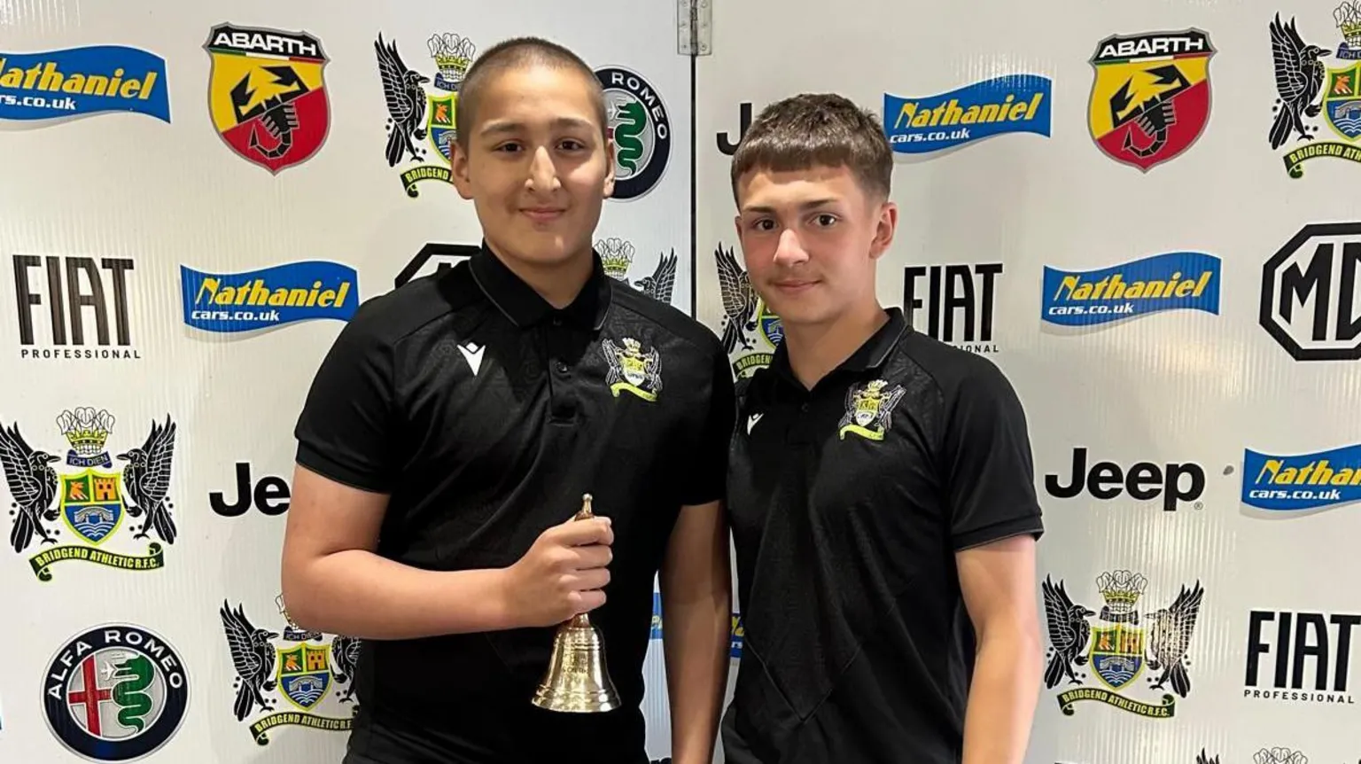 Lloyd Griffiths Ethan in a club rugby shirt with a fellow player. He is holding a brass bell in one hand.