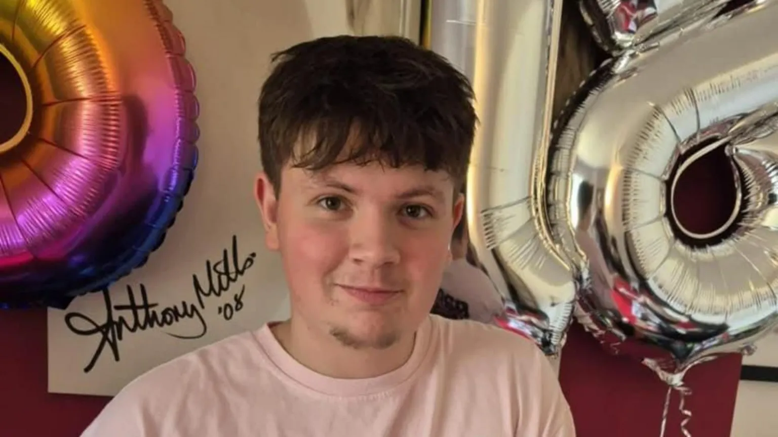 Mills family Picture of young man smiling at the camera. He has dark brown eyes and brown hair with a fringe. He wears a pale coloured t-shirt. There are colourful, celebratory balloons behind him. 
