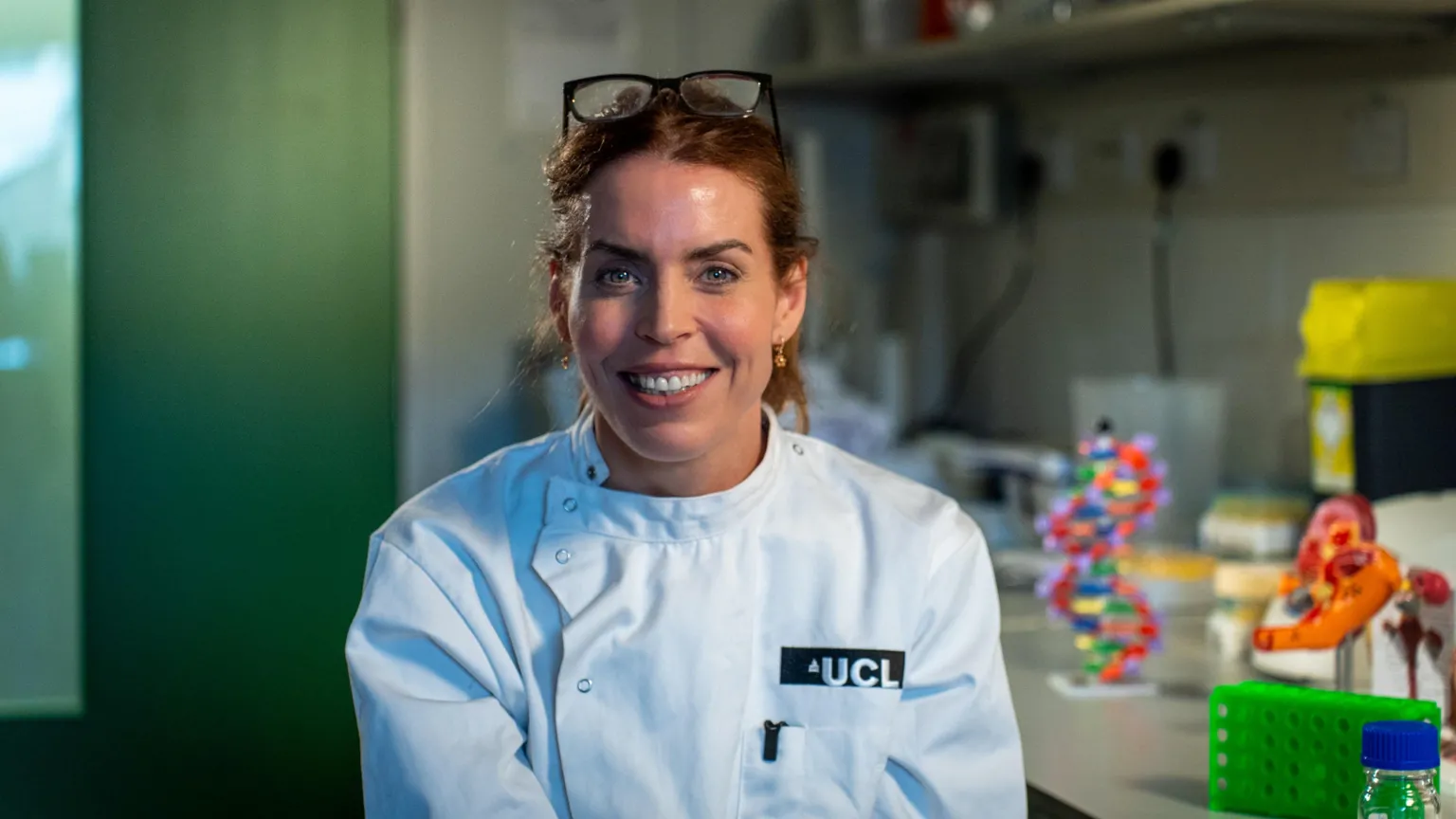 The image shows a person positioned in the foreground of what appears to be a laboratory or research workspace. The person is wearing a white lab coat with visible text on the chest reading “UCL,” and a pair of glasses is resting on top of the person’s head. The background contains various scientific items, including a colourful DNA double-helix model, containers, and laboratory equipment arranged on a work surface. On the right side of the image there are bright objects such as a green rack and a container with a yellow lid, adding contrast to the otherwise neutral environment. 