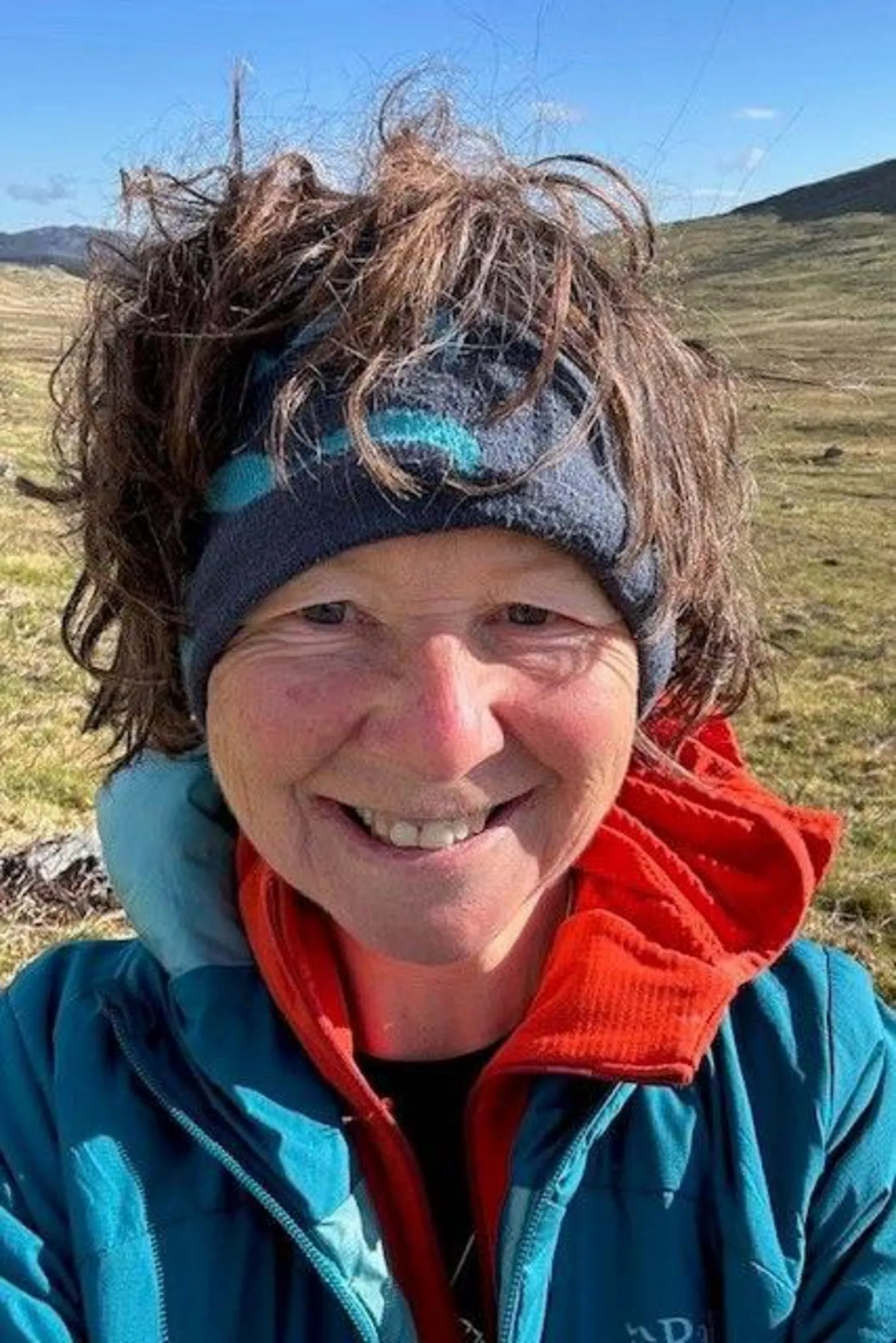 Lorraine McCall A woman is standing in the sunshine under a clear sky. She is smiling and is wearing a blue and red jacket and has a dark blue band holding back her brown hair.