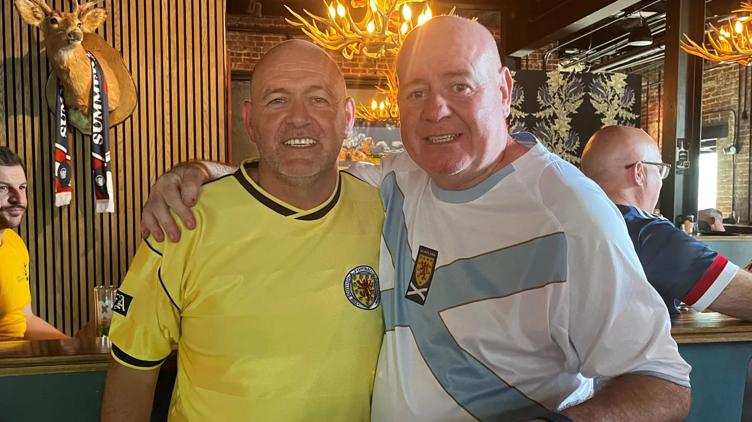 Two people in football shirts standing with arms around each other in a lively bar with antler-style chandeliers, a mounted deer head, and other patrons in the background